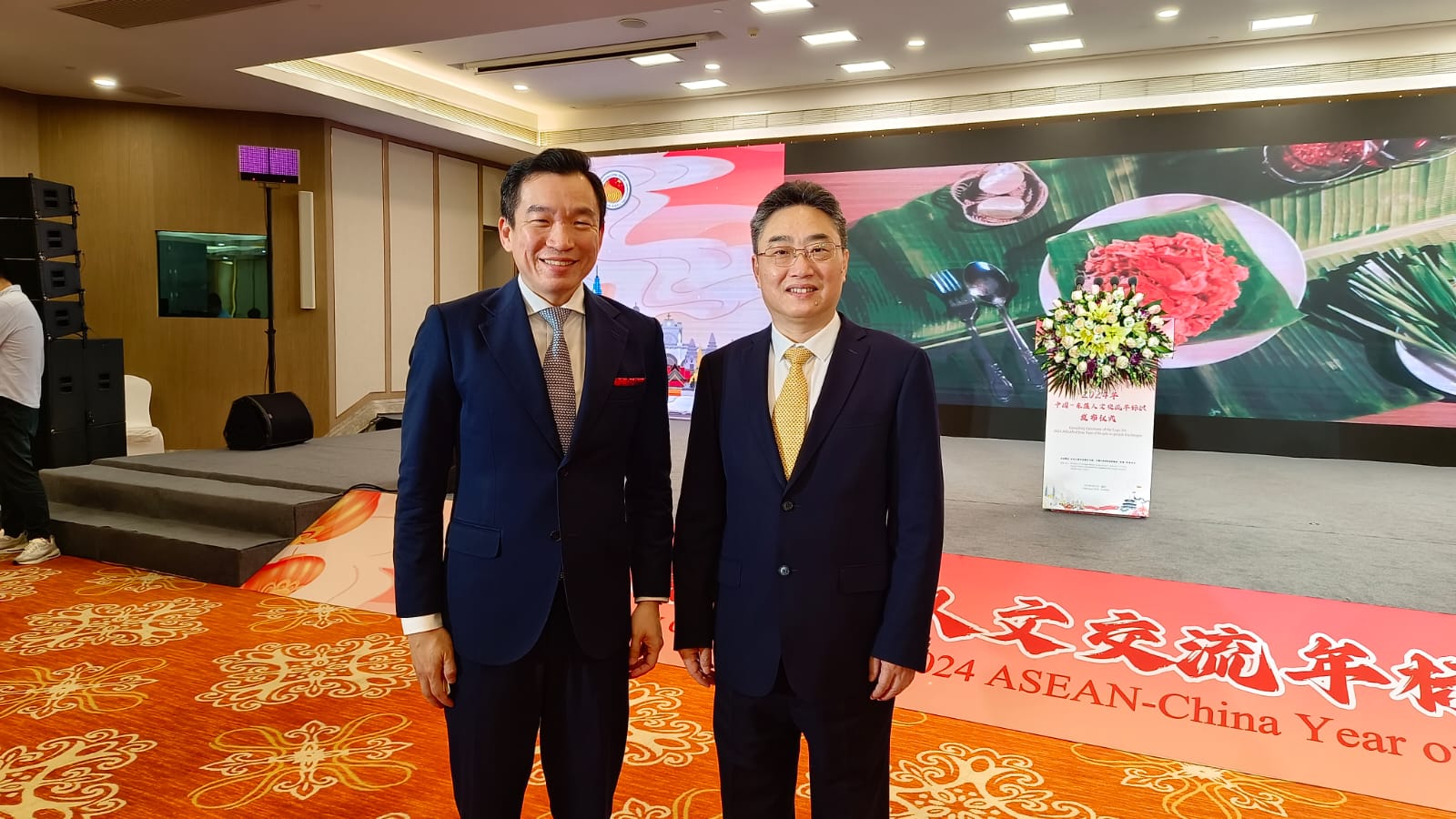 Two men in suits stand smiling in front of a stage with a food image and Chinese lettering.